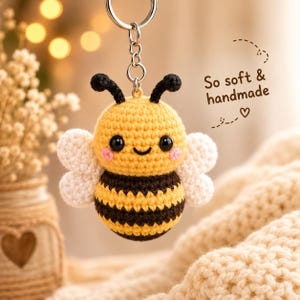 Può includere: Un portachiavi a forma di ape all'uncinetto con corpo a righe gialle e marroni, ali bianche, antenne nere e occhi neri con guance rosa. Il testo "So soft & handmade" con un'icona a forma di cuore è stampato a destra dell'ape.