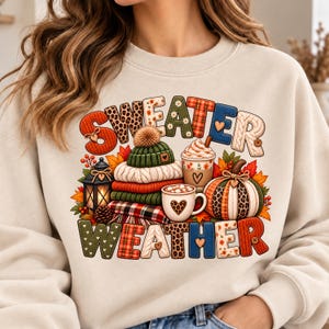 May include: A beige crewneck sweatshirt featuring the text SWEATER WEATHER in colorful patterned letters. The graphic includes a stack of folded sweaters, a green beanie, a lantern, a pumpkin, and mugs of hot cocoa surrounded by autumn leaves.