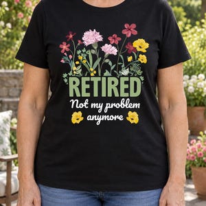 May include: A black short-sleeved t-shirt featuring a colourful floral graphic above the text "RETIRED" in green, followed by the white text "Not my problem anymore" and two small yellow flowers on either side of the final word.