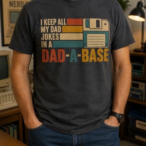 May include: A charcoal gray short-sleeve t-shirt featuring a graphic of a vintage floppy disk and the text I KEEP ALL MY DAD JOKES IN A DAD-A-BASE. The design uses horizontal stripes in red, yellow, and teal colors with bold lettering.