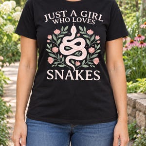 Könnte beinhalten: Ein schwarzes kurzärmeliges T-Shirt mit einem Grafikdruck einer hellrosa Schlange, die sich in der Mitte windet, umgeben von rosa Blumen und grünen Blättern. Der Text "JUST A GIRL WHO LOVES SNAKES" ist in Hellrosa über und unter der Schlange gedruckt.