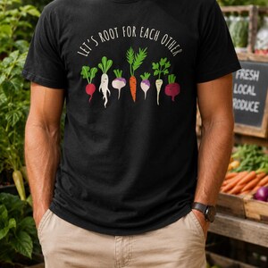 May include: A black short-sleeved t-shirt featuring a graphic of various colourful root vegetables including a radish, carrot, and beet. Above the vegetables, the text reads "LET'S ROOT FOR EACH OTHER" in a white, clean, sans-serif font.