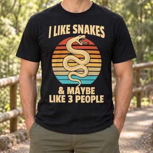 May include: A black short-sleeved t-shirt featuring a graphic of a coiled snake in front of a multi-coloured sunset circle. The text reads I LIKE SNAKES & MAYBE LIKE 3 PEOPLE in a bold, light-coloured font centred on the chest of the garment.