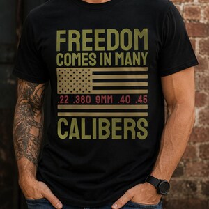 May include: A black short-sleeved t-shirt featuring the text FREEDOM COMES IN MANY CALIBERS in olive green. Below the text is a graphic of an American flag with the numbers .22, .380, 9MM, .40, and .45 printed in red across the stripes.