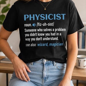 May include: A black short-sleeved t-shirt featuring the text PHYSICIST in blue, followed by the definition: noun. /fiz-uh-sist/ Someone who solves a problem you didn't know you had in a way you don't understand. see also: wizard, magician in white.