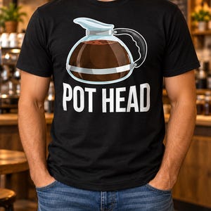Op de afbeelding: Een zwart T-shirt met korte mouwen en een afbeelding van een glazen koffiepot gevuld met donkerbruine koffie. Onder de koffiepot staat de tekst POT HEAD in grote, dikgedrukte, witte hoofdletters in het midden op de borst gedrukt.