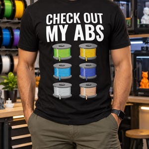 Peut inclure: Un t-shirt noir avec le texte blanc "CHECK OUT MY ABS" au-dessus d'un graphique représentant six bobines de filament pour imprimante 3D disposées en deux colonnes. Les bobines sont vert citron, jaune, bleu clair, bleu foncé, blanc et orange clair.