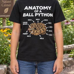 May include: A black short-sleeve t-shirt featuring a graphic of a coiled brown and tan ball python. The text reads ANATOMY OF A BALL PYTHON with arrows pointing to various parts labeled NECK, VISION BEANS, and PBBBBT in white capital letters.