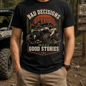 Könnte beinhalten: Ein schwarzes T-Shirt mit einem Grafikdruck eines Geländewagens in einer bergigen Waldlandschaft. Der Text BAD DECISIONS ist über dem Fahrzeug gewölbt und MAKE GOOD STORIES ist darunter in einer fetten, beigefarbenen Schrift gedruckt.