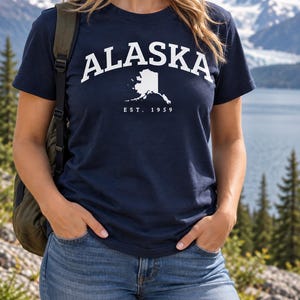 May include: A navy blue short-sleeved t-shirt featuring the white text ALASKA in an arched font above a white silhouette map of the state of Alaska, with the text EST. 1959 printed in white directly below the map graphic on the front of the shirt.