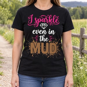 Könnte beinhalten: Ein schwarzes Kurzarm-T-Shirt mit dem rosa Text I sparkle, einem weißen Diamantsymbol, dem weißen Text even in the und dem braunen Text MUD mit einem braunen Schlammspritzer-Grafikdesign hinter dem Wort MUD auf der Vorderseite.