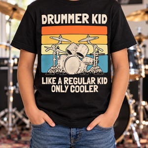 Op de afbeelding: Een zwart t-shirt met korte mouwen en een afbeelding van een drumstel tegen een retro-achtergrond met oranje, gele en blauwe strepen. De tekst luidt DRUMMER KID bovenaan en LIKE A REGULAR KID ONLY COOLER onderaan in witte letters.