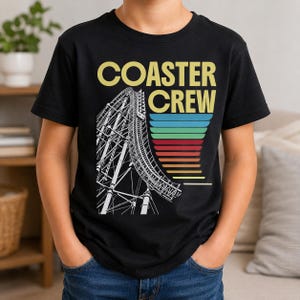 Puede incluir: Una camiseta negra de manga corta con el texto "COASTER CREW" en amarillo. Debajo del texto hay una ilustración de líneas blancas de una vía de montaña rusa y una serie de barras horizontales en colores azul, verde, rojo, naranja y amarillo.