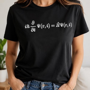 May include: A black short-sleeved t-shirt featuring the white printed Schrödinger equation, iħ ∂/∂t Ψ(r,t) = ĤΨ(r,t), centered on the chest. The garment has a classic crew neckline and a standard fit, paired with blue denim jeans.