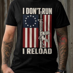 May include: A black short-sleeve t-shirt featuring a graphic print of an American flag with a circle of thirteen stars. A rifle is positioned vertically over the flag. The text I DON'T RUN is printed above and I RELOAD is printed below the graphic.