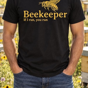 Può includere: Una t-shirt nera a maniche corte con una grafica gialla di un'ape sopra il testo "Beekeeper" e la frase "If I run, you run" stampati in lettere gialle sulla parte del torace del capo di abbigliamento.