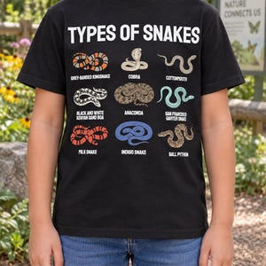 May include: A black short-sleeved t-shirt featuring the white text TYPES OF SNAKES at the top. Below are nine illustrations of snakes with their names: Grey-Banded Kingsnake, Cobra, Cottonmouth, Black and White Kenyan Sand Boa, Anaconda, San Francisco Garter Snake, Milk Snake, Indigo Snake, and Ball Python.