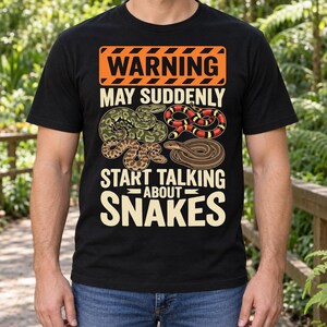 Puede incluir: Una camiseta negra de manga corta con un gráfico de señal de advertencia naranja con el texto "WARNING MAY SUDDENLY START TALKING ABOUT SNAKES" e ilustraciones de cuatro serpientes diferentes en colores verde, rojo, amarillo, negro y marrón.