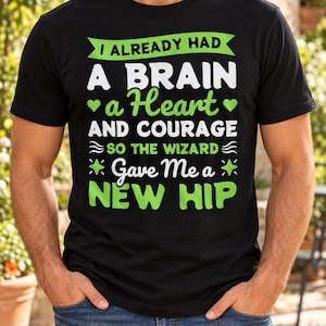 Puede incluir: Una camiseta negra de manga corta con texto en verde y blanco que dice: I ALREADY HAD A BRAIN a Heart AND COURAGE SO THE WIZARD Gave Me a NEW HIP. El diseño incluye pequeños iconos de corazones, ondas y estrellas.