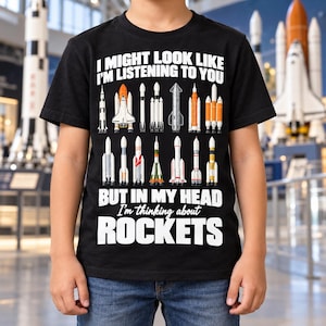 May include: A black short-sleeve t-shirt featuring white text that reads I MIGHT LOOK LIKE I'M LISTENING TO YOU BUT IN MY HEAD I'm thinking about ROCKETS. The design includes a graphic illustration of twelve different space rockets.