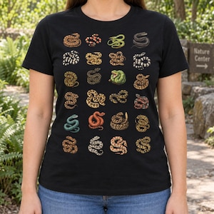 Può includere: Una t-shirt nera a maniche corte con una griglia di ventiquattro illustrazioni di serpenti distinti in vari colori e motivi, tra cui marrone, verde, rosso e nero, disposti in sei righe e quattro colonne sulla parte anteriore del capo.