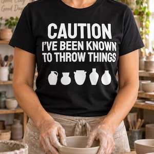 May include: A black short-sleeved t-shirt featuring the white text CAUTION I'VE BEEN KNOWN TO THROW THINGS above a row of five white pottery silhouettes. The shirt has a crew neckline and is made of a solid black fabric with bold white lettering.