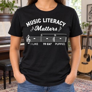 May include: A black short-sleeved t-shirt featuring white text that reads MUSIC LITERACY Matters, a musical staff with a treble clef, and the text I LIKE TO EAT PUPPIES. The design includes musical notes and staff lines printed on the front.