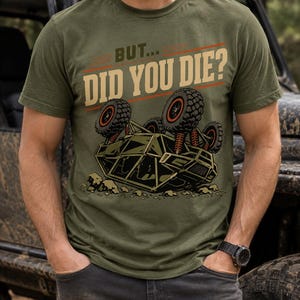 Op de afbeelding: Een olijfgroen T-shirt met korte mouwen en een afbeelding van een omgekanteld terreinvoertuig met grote banden. Boven het voertuig staat de tekst BUT... DID YOU DIE? in een vet, beige schreefloos lettertype met dunne oranje lijnen.