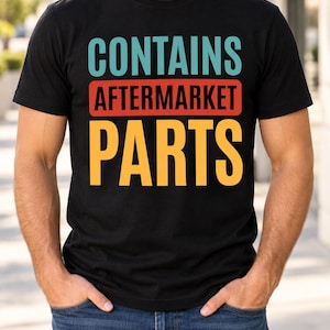 Puede incluir: Una camiseta negra de manga corta con el texto CONTAINS en azul claro, AFTERMARKET en negro sobre un fondo rectangular rojo, y PARTS en amarillo. El gráfico está centrado en el pecho de la camiseta, que se lleva con unos vaqueros azules.