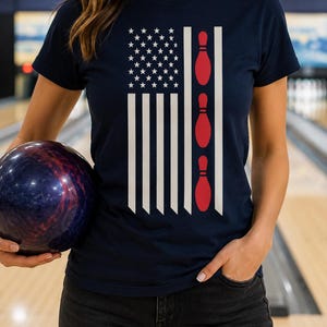 May include: A navy blue short-sleeve t-shirt featuring a graphic design that combines the American flag with three red bowling pins. The design includes white stars and vertical stripes, with the bowling pins replacing one of the stripes.