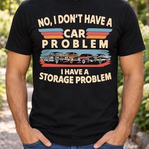 Puede incluir: Una camiseta negra de manga corta con un gráfico que incluye el texto NO, I DON'T HAVE A CAR PROBLEM I HAVE A STORAGE PROBLEM. El diseño muestra cuatro coches clásicos de varios colores sobre franjas horizontales en azul, naranja y rojo.