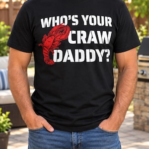 May include: A black short-sleeved t-shirt featuring a red graphic of a crawfish and the white text "WHO'S YOUR CRAW DADDY?" printed on the chest. The shirt has a crew neckline and a standard fit, showcasing a bold design with a playful seafood theme.