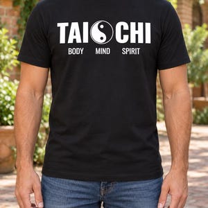 Könnte beinhalten: Ein schwarzes Kurzarm-T-Shirt mit weißem Aufdruck TAI CHI, bei dem ein Yin-Yang-Symbol den Buchstaben I ersetzt. Unter dem Haupttext sind die Wörter BODY MIND SPIRIT in einer kleineren weißen Schrift auf der Brust gedruckt.