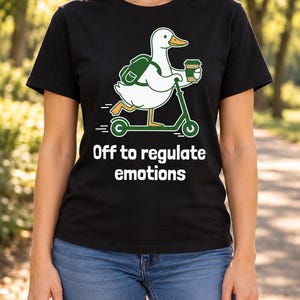 Op de afbeelding: Een zwart T-shirt met korte mouwen en een afbeelding van een witte eend met een oranje snavel, een groene rugzak, rijdend op een groene step en een koffiebeker vasthoudend. Onder de afbeelding staat: Off to regulate emotions.