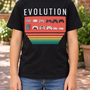 Könnte beinhalten: Ein schwarzes Kurzarm-T-Shirt mit dem Wort EVOLUTION in weißer Schrift über einer Grafik verschiedener Videospiel-Controller, die in zwei Reihen angeordnet sind. Unter den Controllern befindet sich ein Retro-Sonnenuntergangs-Design mit horizontalen Streifen in Rot, Orange, Gelb, Grün und Türkis.