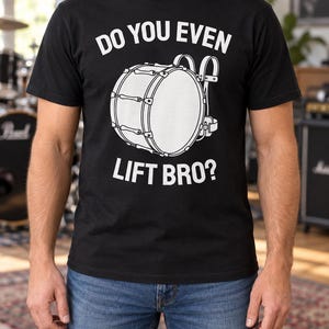 Puede incluir: Una camiseta negra de manga corta con un gráfico blanco de un bombo de marcha con correas. El texto "DO YOU EVEN" está impreso en blanco sobre el tambor, y el texto "LIFT BRO?" está impreso en blanco debajo del gráfico del tambor.