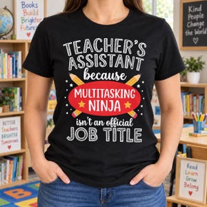 Puede incluir: Una camiseta negra de manga corta con texto en blanco y amarillo que dice Teacher's Assistant because Multitasking Ninja isn't an official Job Title. El diseño incluye dos lápices amarillos cruzados y un elemento de fondo ovalado rojo.