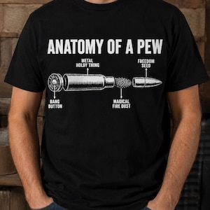 Puede incluir: Una camiseta negra con un diagrama gráfico blanco titulado ANATOMY OF A PEW. La ilustración muestra una bala desmontada con etiquetas que señalan las partes: BANG BUTTON, METAL HOLDY THING, MAGICAL FIRE DUST y FREEDOM SEED.