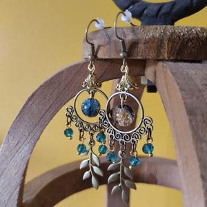 Può includere: Un paio di orecchini pendenti con cerchi in filigrana di metallo color bronzo, ciascuno centrato con una perlina rotonda. La perlina sinistra è blu scuro, quella destra è oro e nera. Piccole perline di cristallo verde acqua e ciondoli a forma di foglia color bronzo pendono dai cerchi.