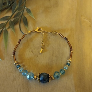 Op de afbeelding: Een kralenarmband met een centrale donkerblauwe gemarmerde kraal, geflankeerd door gefacetteerde lichtblauwe kristallen en goudkleurige tussenstukjes. De band bevat bruine cilindervormige kralen en kleine donkere kralen, afgewerkt met een goudkleurige sluiting.