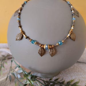 Può includere: Una collana di perline con ciondoli a forma di foglia in bronzo metallizzato, piccole perline rotonde in bronzo, perline bianche e perline di cristallo azzurro sfaccettate, con una disposizione simmetrica di foglie e perline blu.