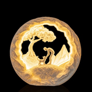 Pode incluir: Uma escultura circular iluminada com aspeto de pedra, apresentando um relevo de uma pessoa com um cão sob uma árvore, perante uma montanha. A peça emite uma luz quente a partir do interior da sua moldura circular texturizada.