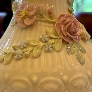 May include: A cream-colored ceramic pitcher featuring raised, three-dimensional floral decorations. The surface is adorned with sculpted pink roses, small light blue blossoms, and green leaves, set against a vertically ribbed and embossed body.