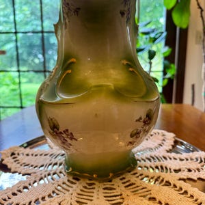 May include: A ceramic jug featuring a cream-coloured base with a gradient green finish at the top and bottom. The vessel is decorated with small painted purple floral clusters and gold-toned trim along the rim and base, with a curved spout.