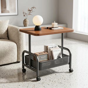 May include: A mobile side table with a rectangular wooden top and a dark gray metal frame. The unit features a lower storage basket containing a small speaker and books. A spherical lamp and a white candle sit on the wooden surface of the table.