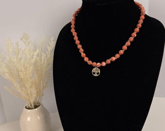 Può includere: Una collana composta da perline di pietra rotonde e sfaccettate color salmone, infilate insieme a piccoli distanziatori color oro. Un ciondolo circolare color oro raffigurante un dettagliato albero della vita pende dal centro.