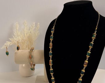 Può includere: Un set di gioielli composto da una collana lunga, un bracciale e un paio di orecchini pendenti. Ogni pezzo presenta perline a forma di botte di colore verde scuro e marrone screziato, separate da piccoli distanziatori in metallo dorato.