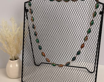 Può includere: Una collana lunga caratterizzata da perline ovali alternate in tonalità verde smeraldo intenso e marrone rame maculato. Le perline sono collegate da sottili maglie metalliche, creando un filo continuo esposto su un supporto in rete metallica nera.
