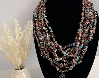 Può includere: Una collana a più fili composta da numerose piccole pietre preziose irregolari e levigate nei toni del turchese, rosa, marrone, nero e bianco. La collana presenta un ciondolo centrale in metallo argentato con un piccolo inserto turchese.
