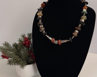 Può includere: Una collana caratterizzata da una serie di pietre multicolori di forma irregolare alternate a perline in metallo argentato testurizzato. Il design include un elemento centrale allungato e affusolato in metallo argentato, affiancato da due piccole pietre arancioni.